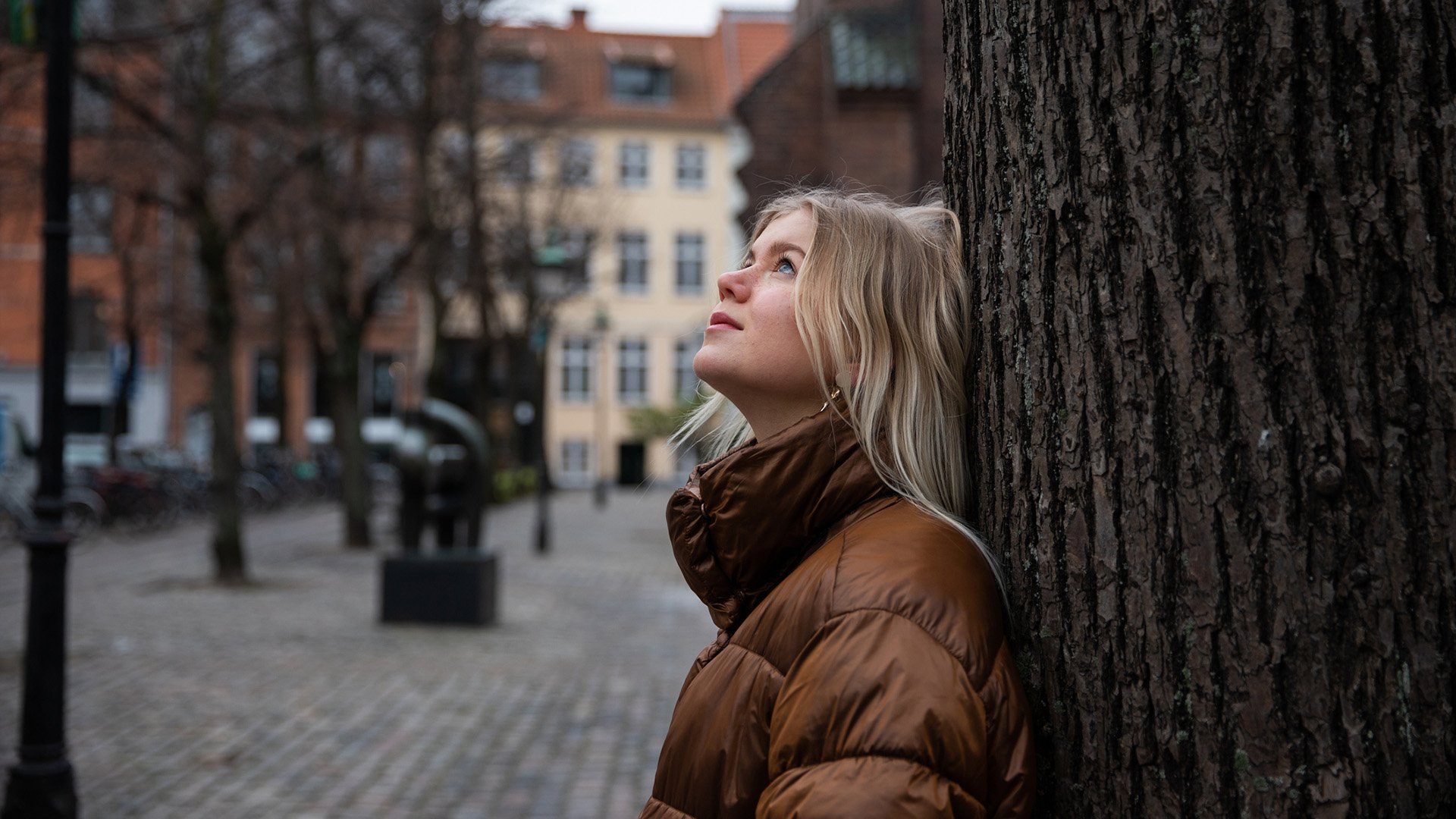 En person med långt blont hår står lutad mot ett träd i en stadsmiljö.